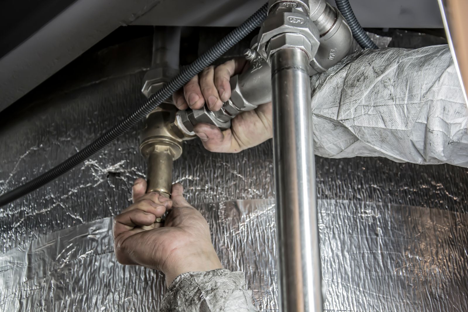 Two working hands in a grey protective sleeve fitting a brass valve onto steel and chrome pipework against silver insulation foil.