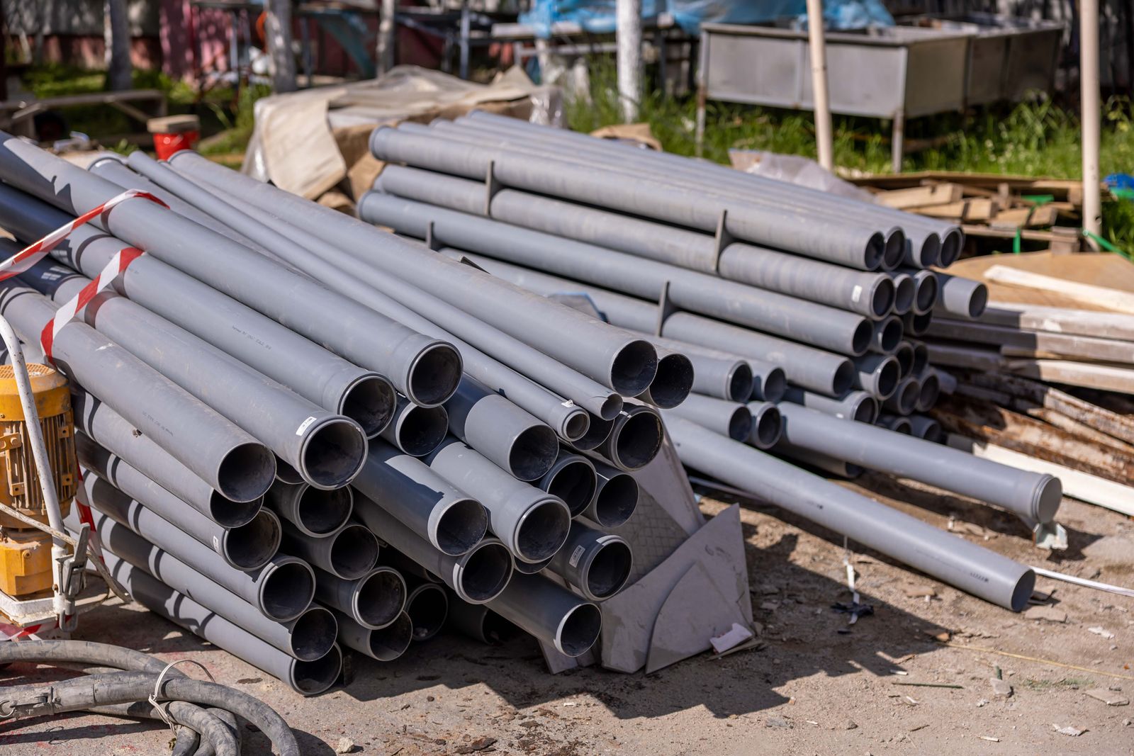Two stacks of grey PVC drainage pipes lying on bare ground at a construction site, with red-and-white tape on one stack, a yellow machine to the left and timber and a tarpaulin behind.